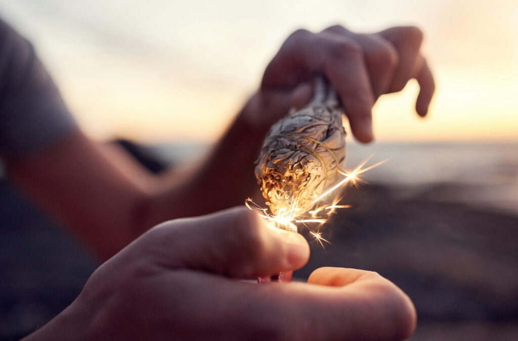 Hand, flame and sage burning at the beach for a spiritual ritual and meditation practice. Herb, fin